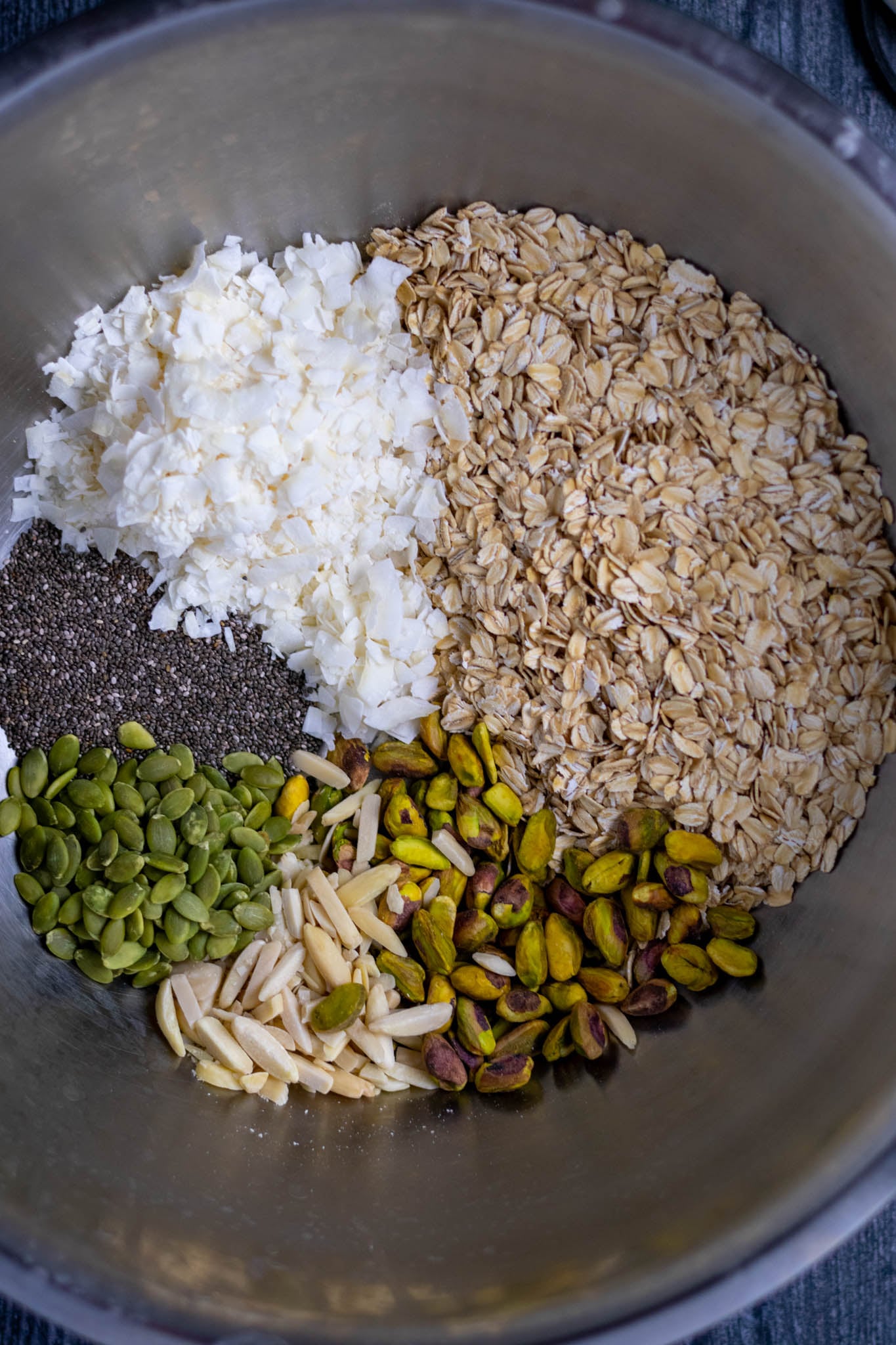 Overhead of large mixing bowl with dry ingredients including coconut, oats, chia, pumpkin seeds, pistachios, and almonds