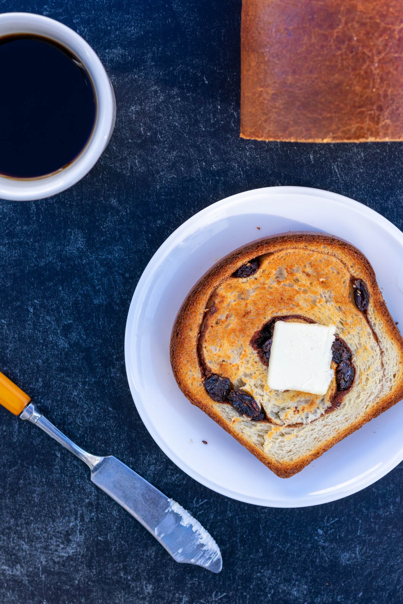 Overhead of toasted sourdough cinnamon raisin bread on plate with slab of butter and coffee