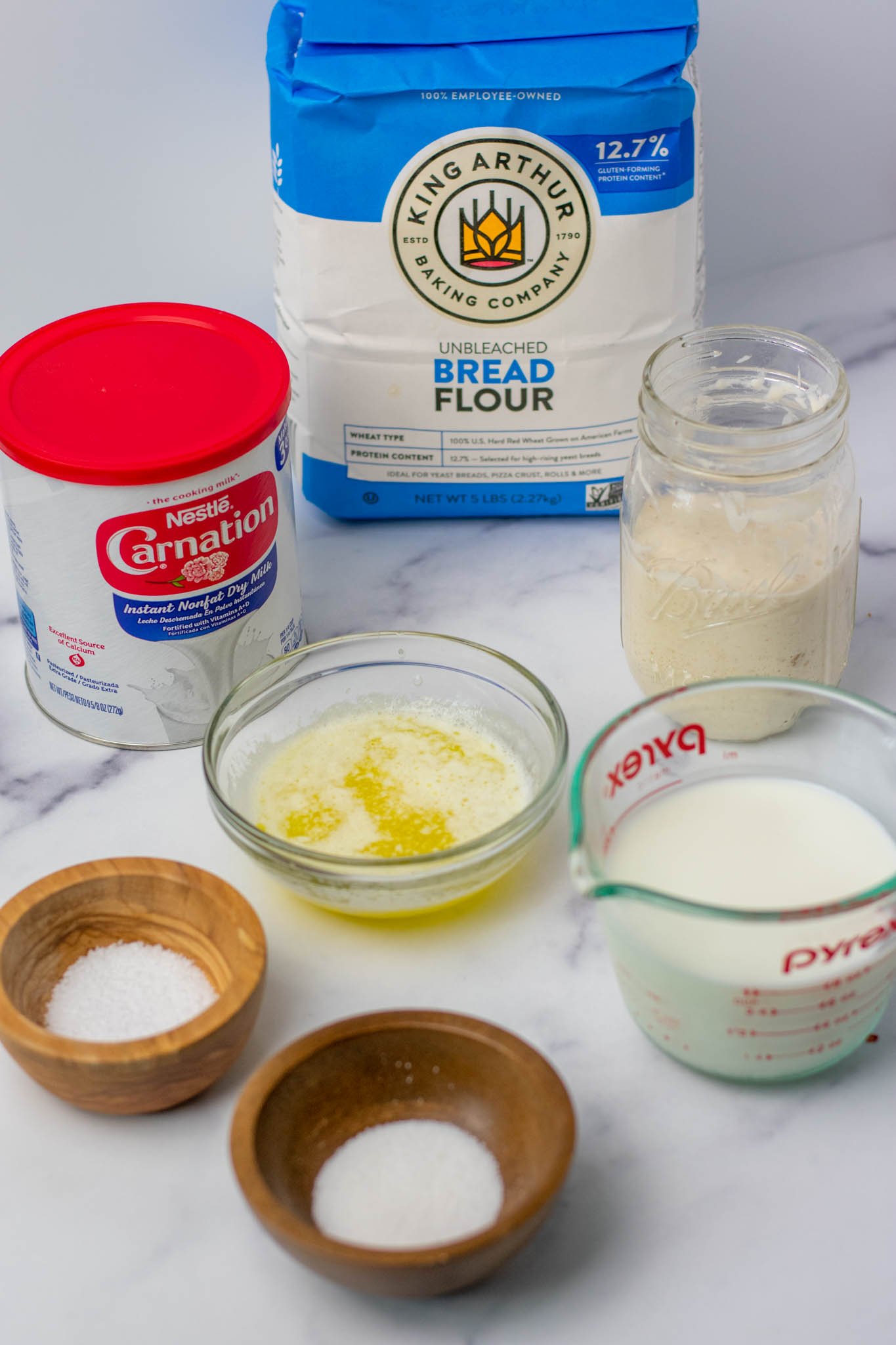Ingredients needed for sourdough sandwich bread including flour, dried milk powder, levain, butter, salt, sugar, and milk