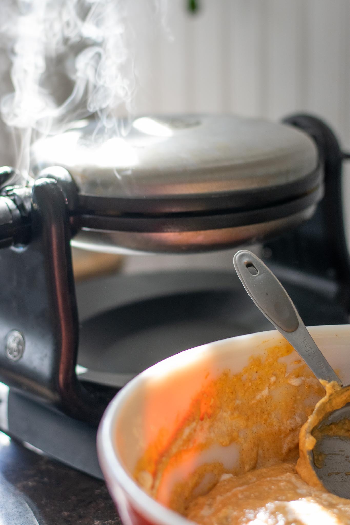 Waffle iron steaming in background with Sourdough Pumpkin Waffle batter in bowl