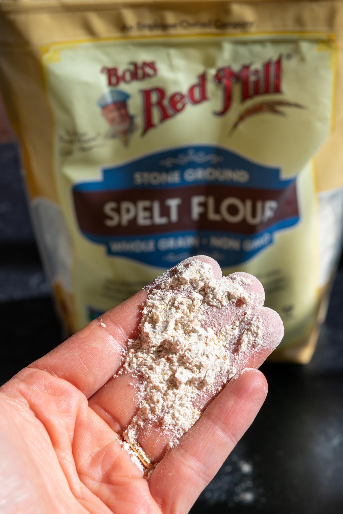 Hand holding spelt flour with bag of spelt flour in background