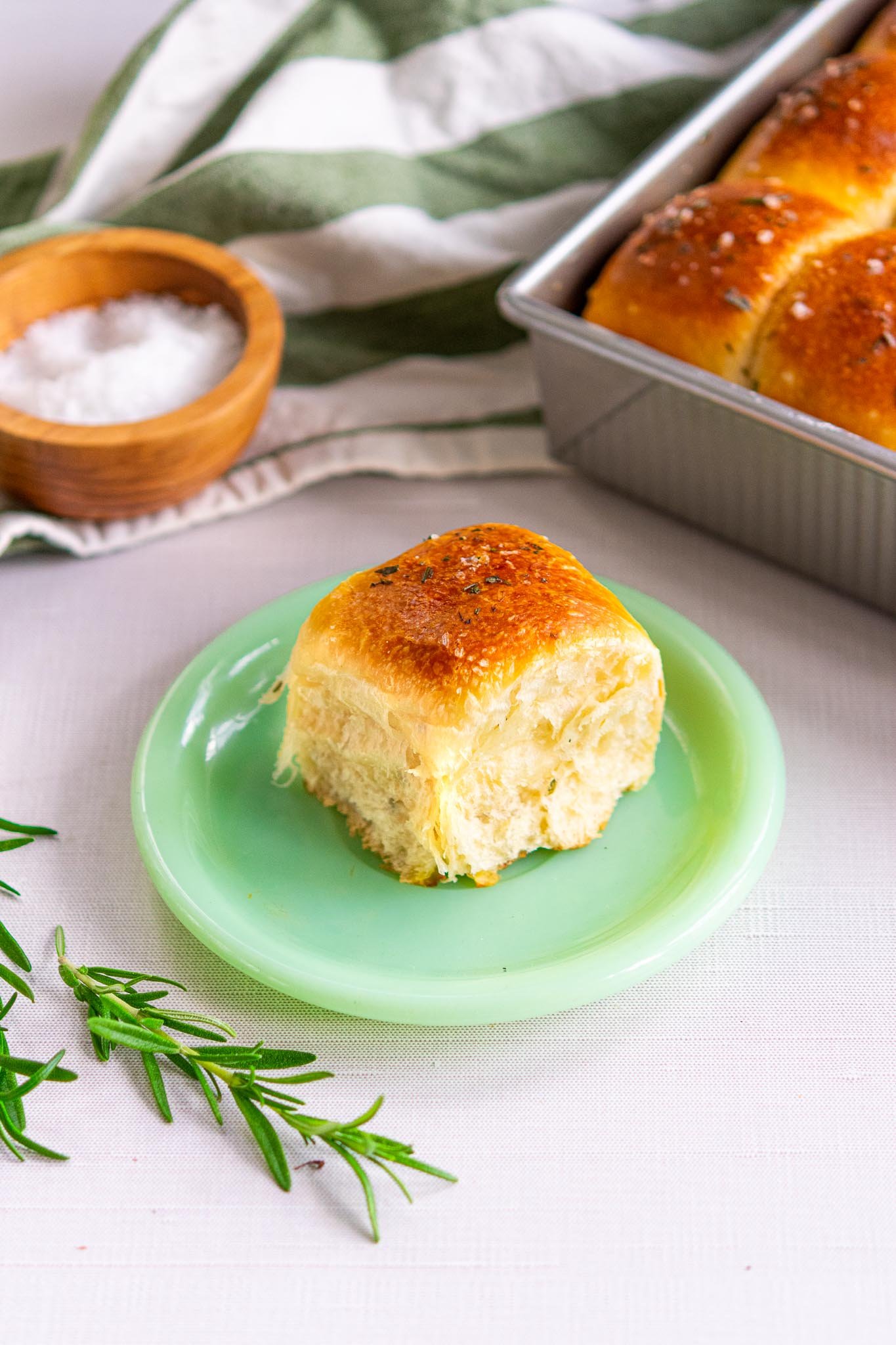 One sourdough dinner roll with rosemary on a plate with sprigs of rosemary and a small bowl of flaky salt