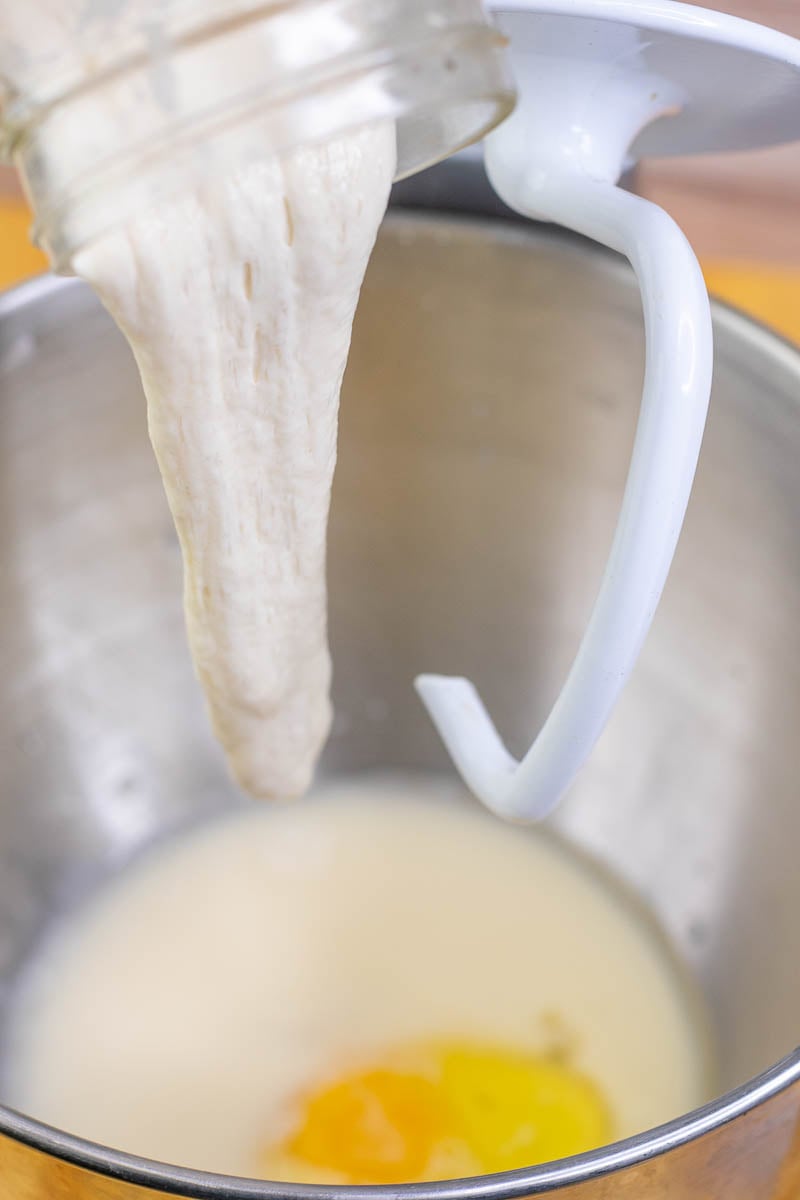 Pouring sourdough starter from a jar into the bowl of a stand mixer with the bread hook attachment