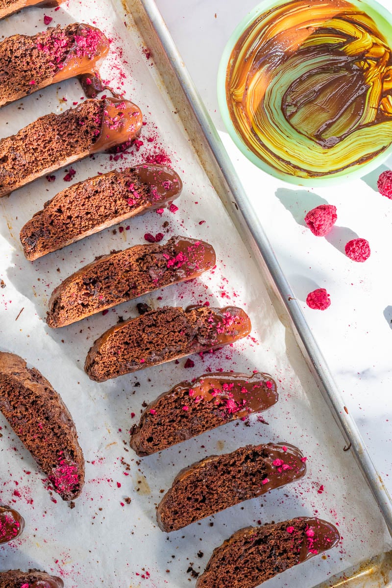 Chocolate raspberry sourdough biscotti on a baking sheet with a bowl of melted chocolate