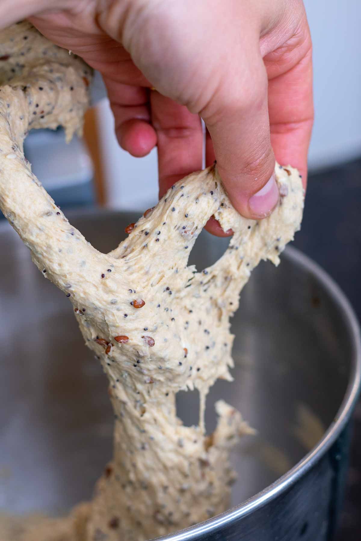 Hand pulling a piece of seeded whole wheat sourdough bread dough and it tearing