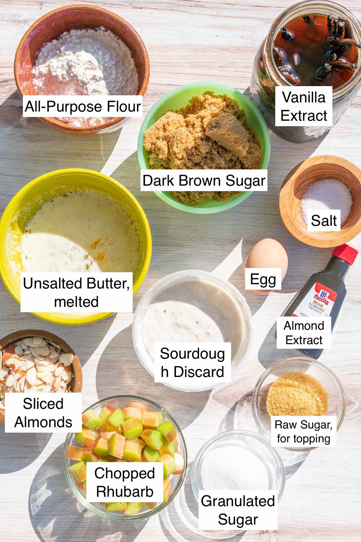 Labeled ingredients needed for Sourdough blondies with rhubarb and almonds including flour, butter, vanilla, salt, egg, almond extract, sourdough discard, almonds, rhubarb, sugar, raw sugar, and dark brown sugar.