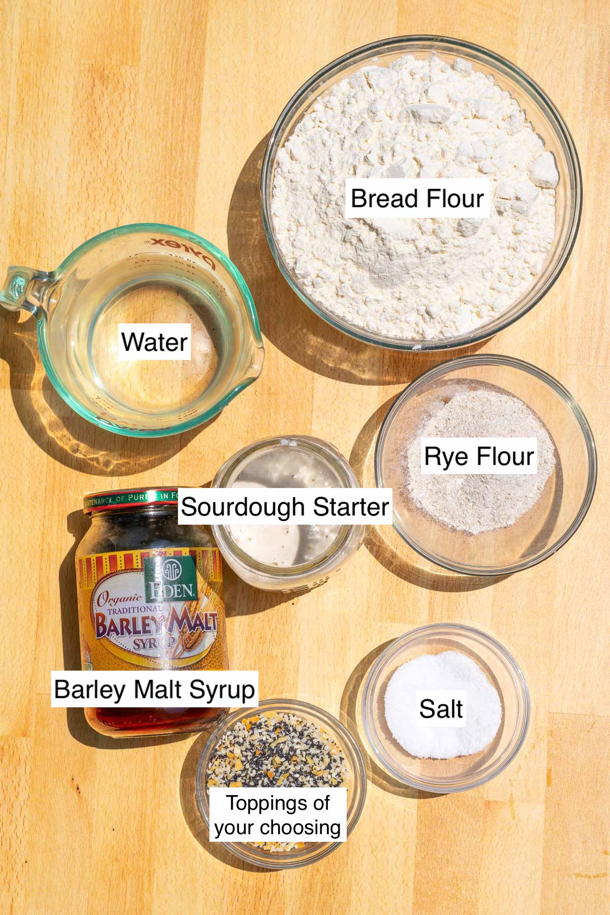 Ingredients needed for sourdough bagels including bowls of bread flour, rye flour, salt, sourdough starter, water, barley malt syrup, and toppings
