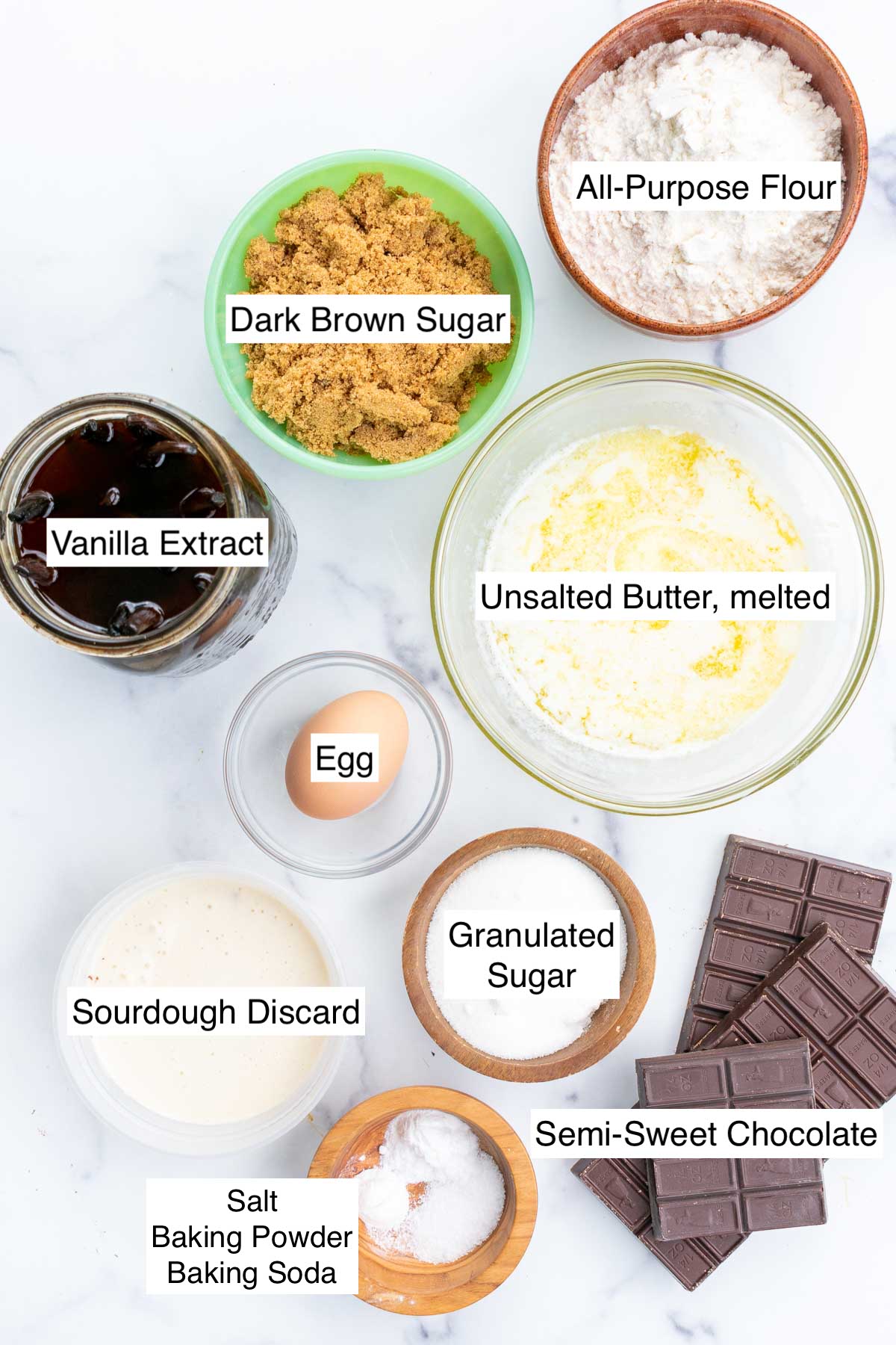 Bowls of ingredients needed for Sourdough discard chocolate chip cookies including all-purpose flour, melted butter, dark brown sugar, granulated sugar, vanilla extract, sourdough discard, an egg, chocolate, baking powder, baking soda, and salt.