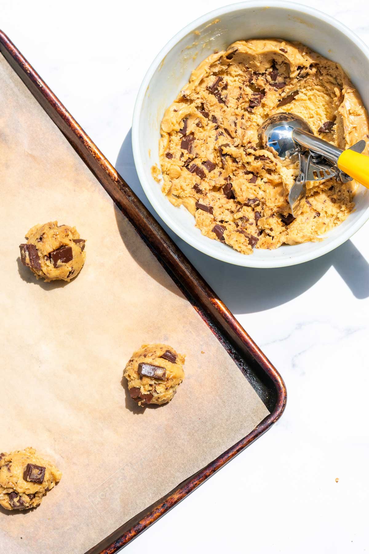 Bowl of chocolate chip cookie dough with a cookie scoop and a baking sheet with cookie balls.