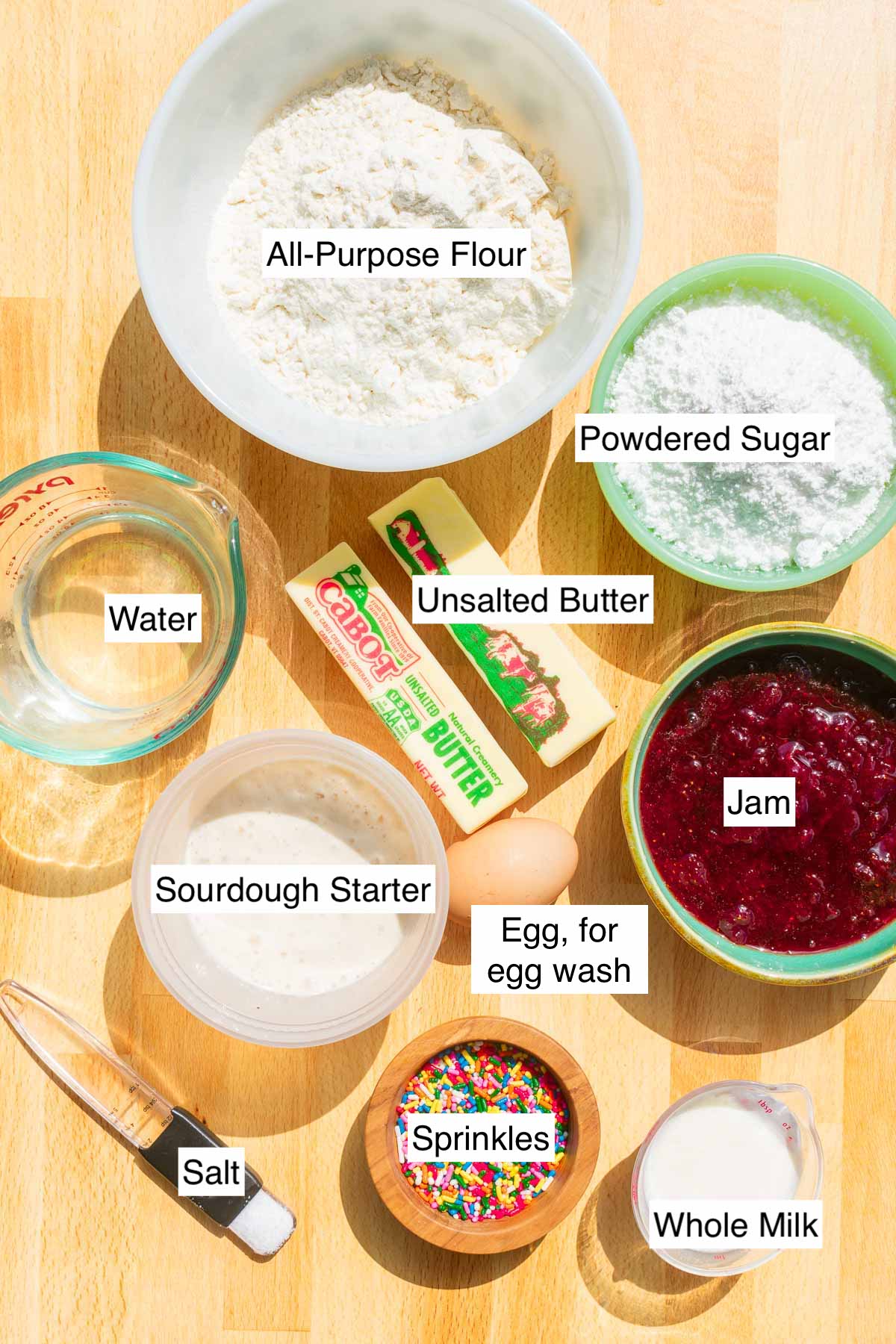 Ingredients needed for sourdough pop tarts including all-purpose flour, granulated and powdered sugar, jam, salt, water, sourdough discard, unsalted butter, sprinkles, milk, and an egg.