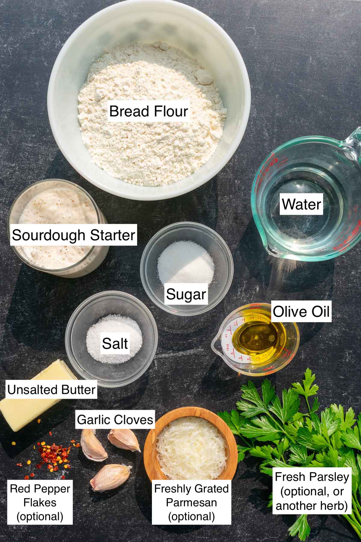 Labeled ingredients needed for sourdough garlic knots including bread flour, water, olive oil, sourdough starter, red pepper flakes, garlic cloves, parsley, parmesan, sugar, salt, and unsalted butter