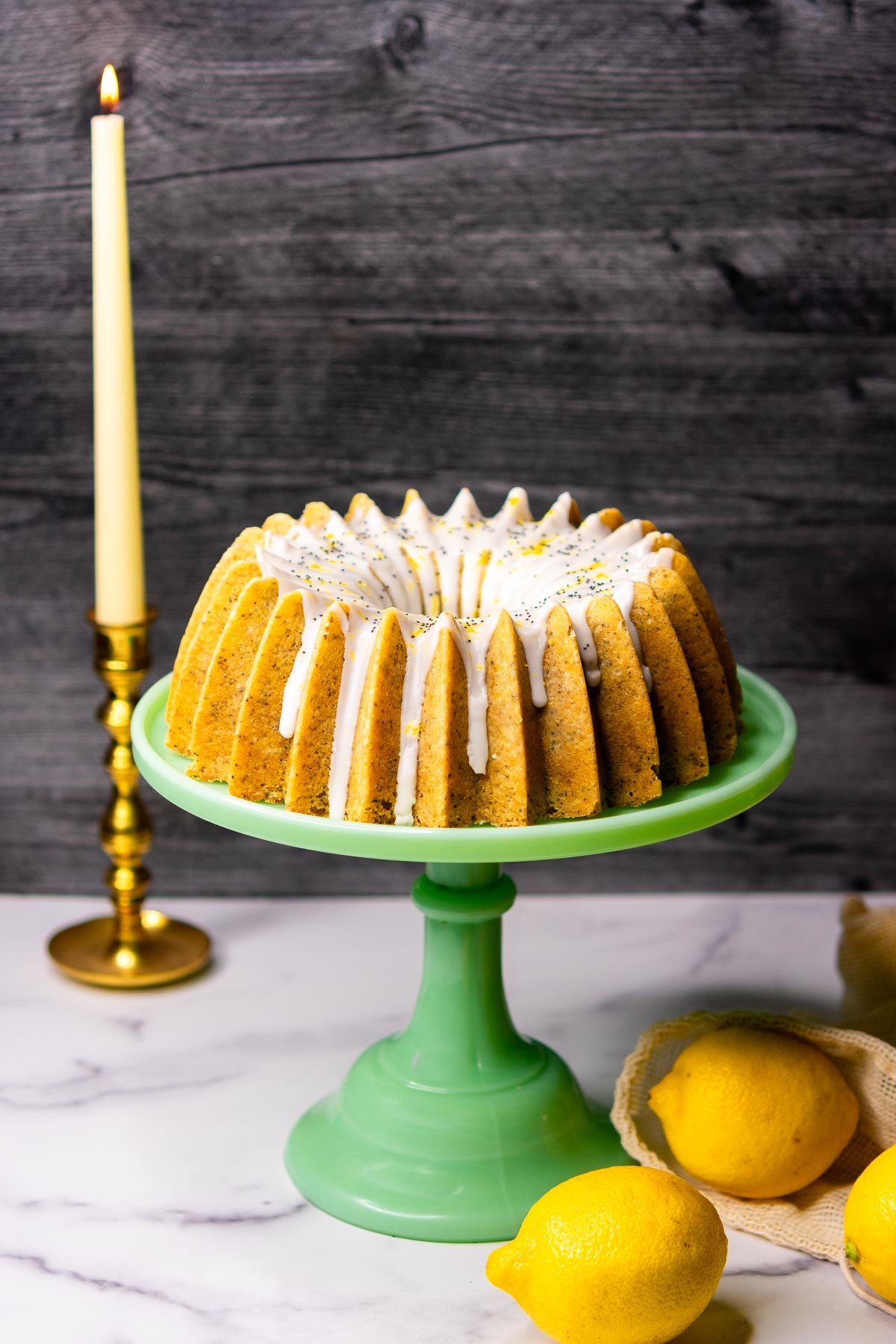 Sourdough lemon poppy seed bundt cake on a cake stand with a candle in the background and a bag of lemons