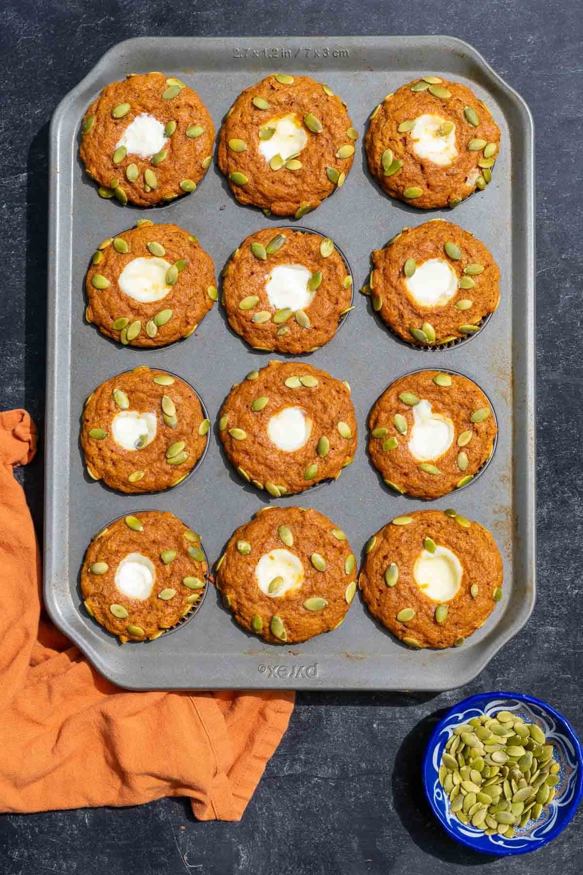 Baked sourdough pumpkin cream cheese muffins in muffin pan with a bowl of pumpkin seeds and an orange towel.