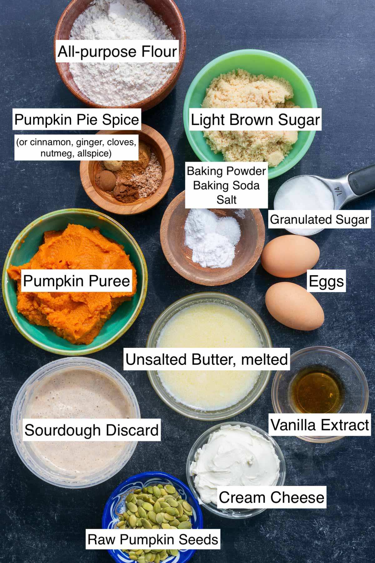 Labeled ingredients needed for sourdough pumpkin cream cheese muffins bowls of all-purpose flour, sourdough discard, light brown sugar, granulated sugar, eggs, unsalted melted butter, pumpkin seeds, pumpkin puree, salt, baking soda, baking powder, vanilla extract, cream cheese, and pumpkin pie spices.