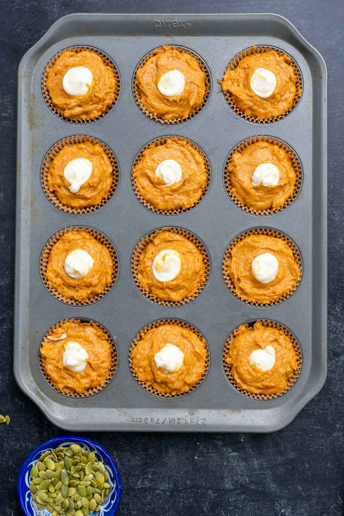 Twelve sourdough pumpkin cream cheese muffins in a muffin pan with a bowl of pumpkin seeds to the side.