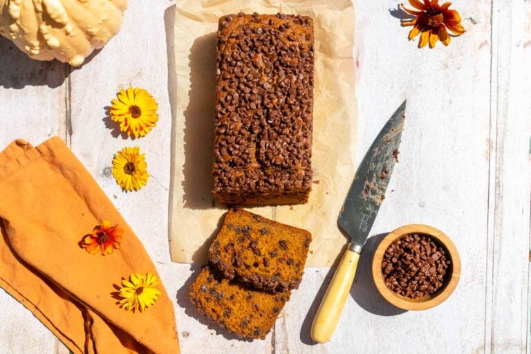 Sourdough pumpkin chocolate chip bread sliced with an orange towel, flowers, and bowl of chocolate chips.