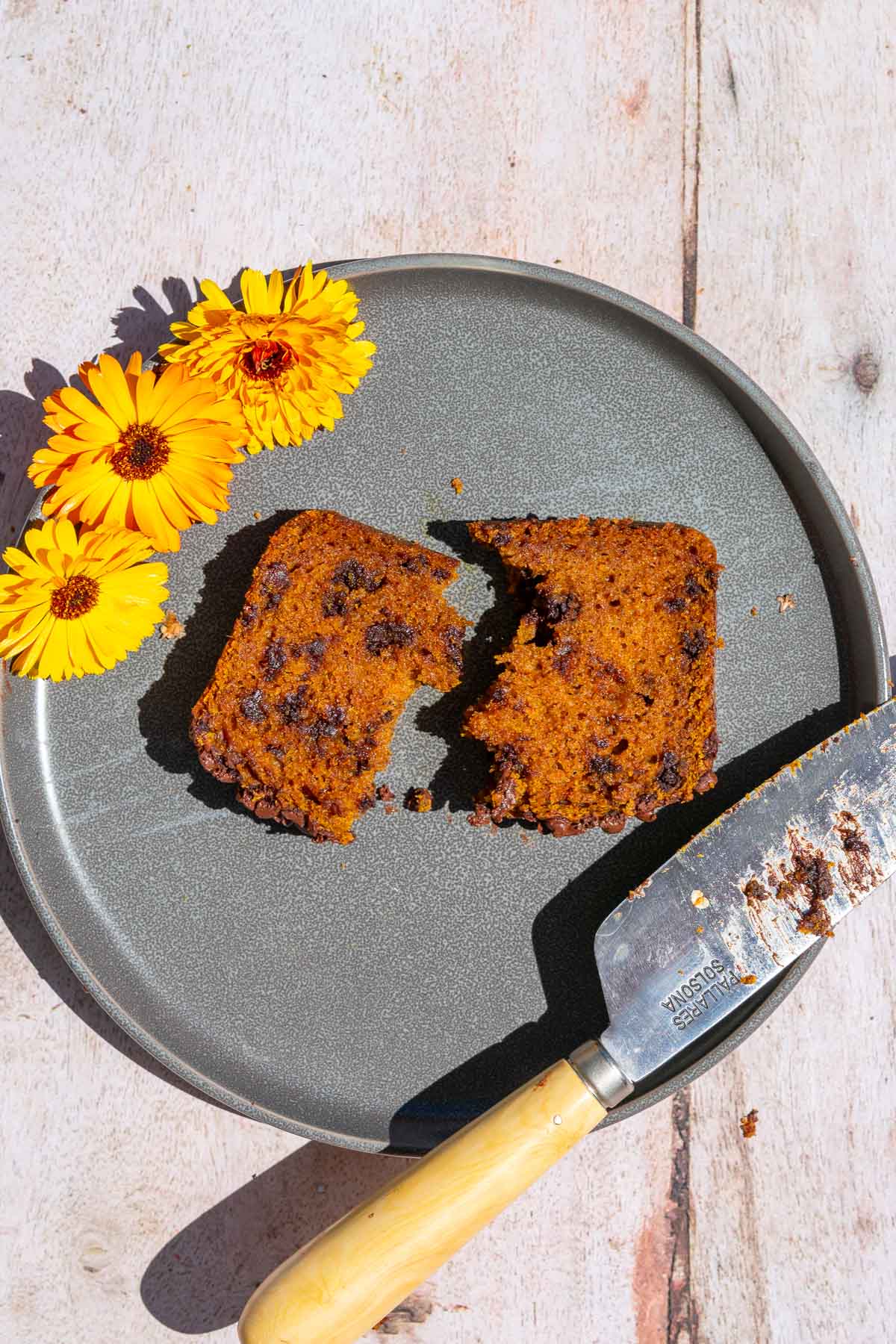 Slice of sourdough pumpkin chocolate chip bread on a plate brokwne in half with a knife and flowers.
