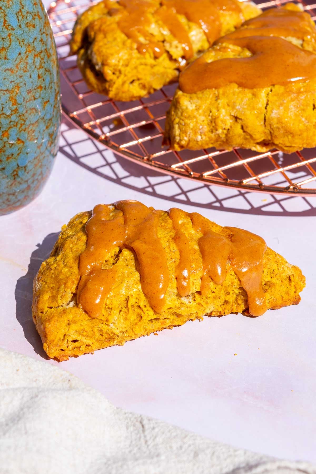 A sourdough pumpkin scone with pumpkin glaze with a towel, flower vase, and more scones in the background.