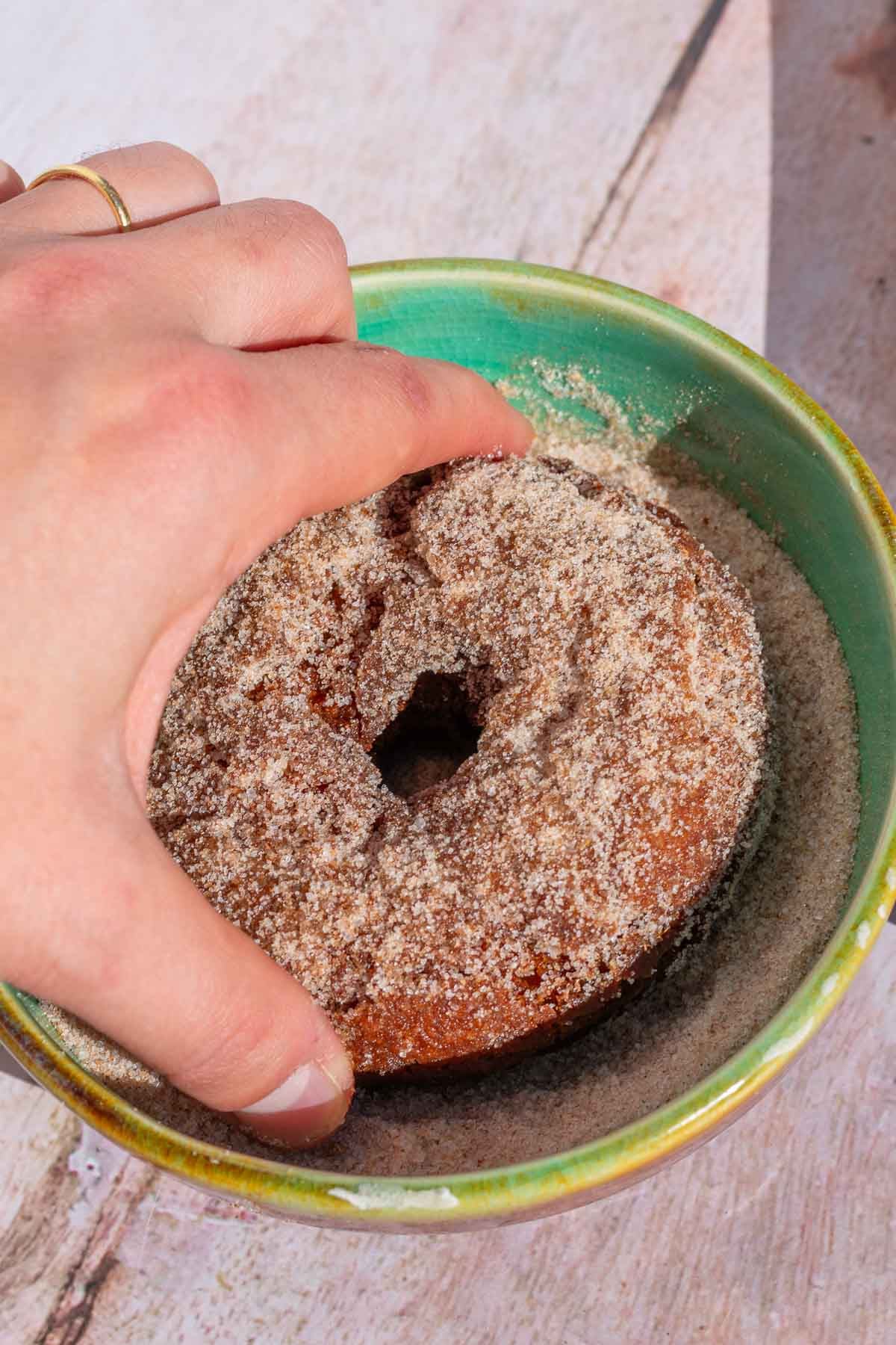 Hand dipping a sourdough apple cider donut in a bowl of cinnamon sugar.