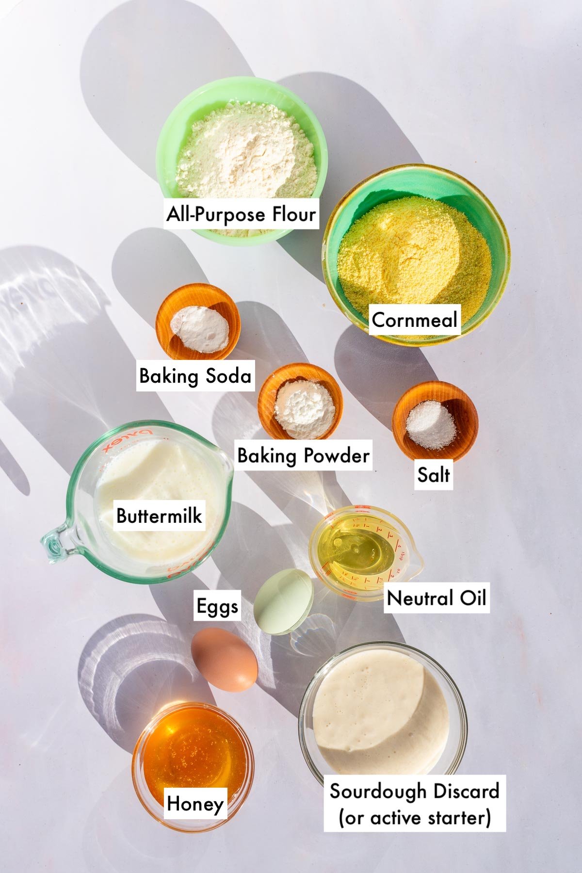 Labeled bowls of ingredients needed for sourdough corn muffins including all-purpose flour, cornmeal, baking soda, baking powder, salt, buttermilk, honey, neutral oil, eggs, and sourdough discard.