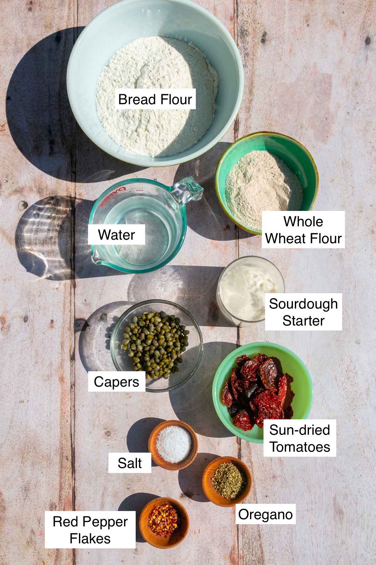 Bowls of labeled ingredients needed to make sun-dried tomato sourdough bread including bread flour, whole wheat flour, water, capers, sun-dried tomatoes, sourdough  starter, salt, oregano, and red pepper flakes.
