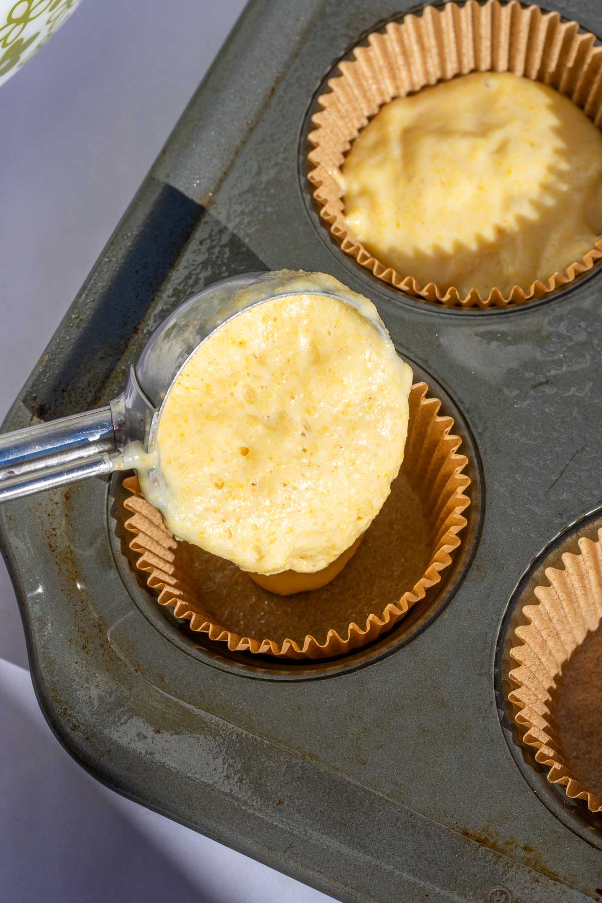 Scooping sourdough corn muffin into a lined muffin pan with a cookie scoop.