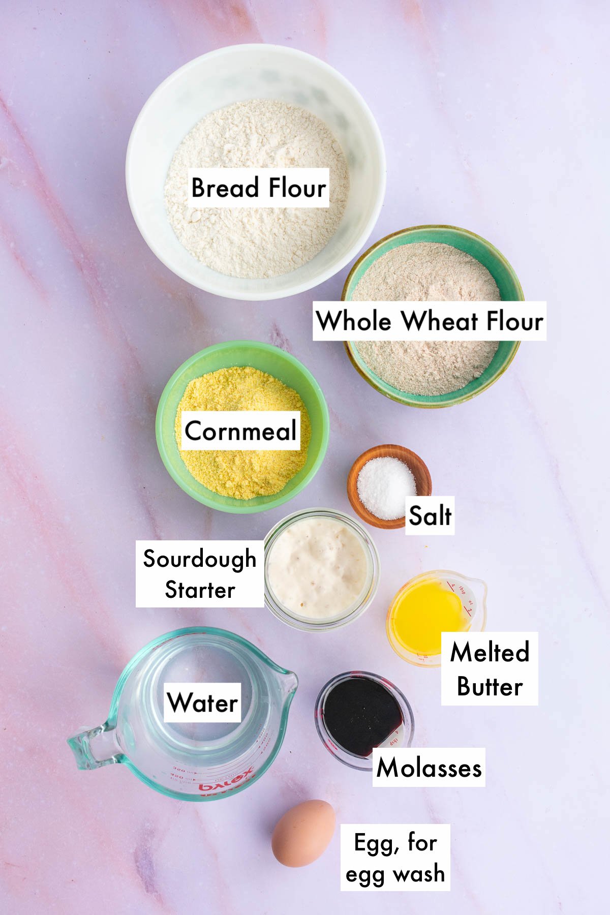 Bowls of labeled ingredients needed for sourdough anadama bread including bread flour, whole wheat  flour, cornmeal, salt, molasses, sourdough starter, melted butter, water, and an egg for egg wash.