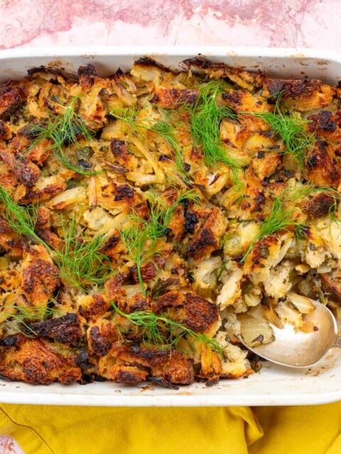 Sourdough stuffing with fennel in a baking dish with a spoon scooping some and fresh herbs.