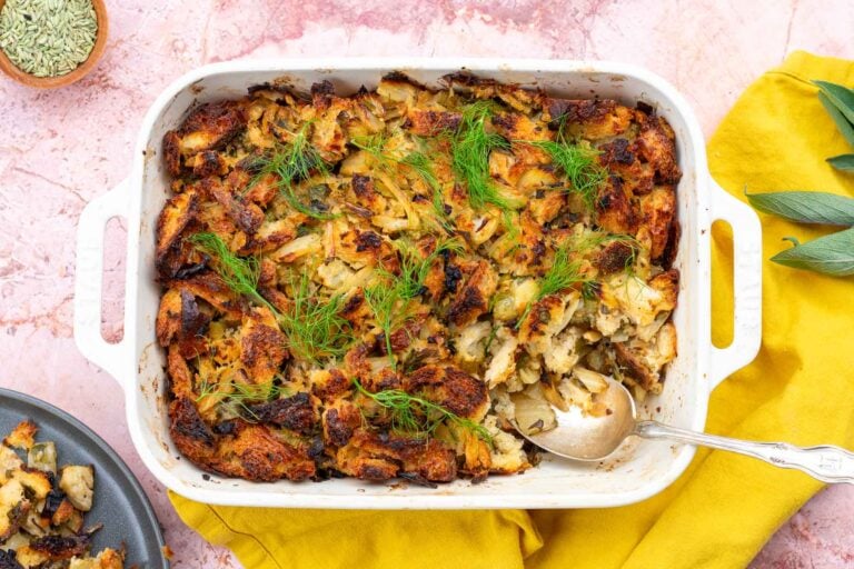 Sourdough stuffing with fennel in a baking dish with a spoon scooping some and fresh herbs.
