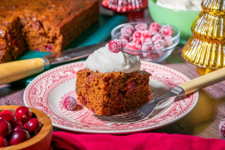 A slice of sourdough gingerbread cake with whipped cream and sugared cranberries on a plate.