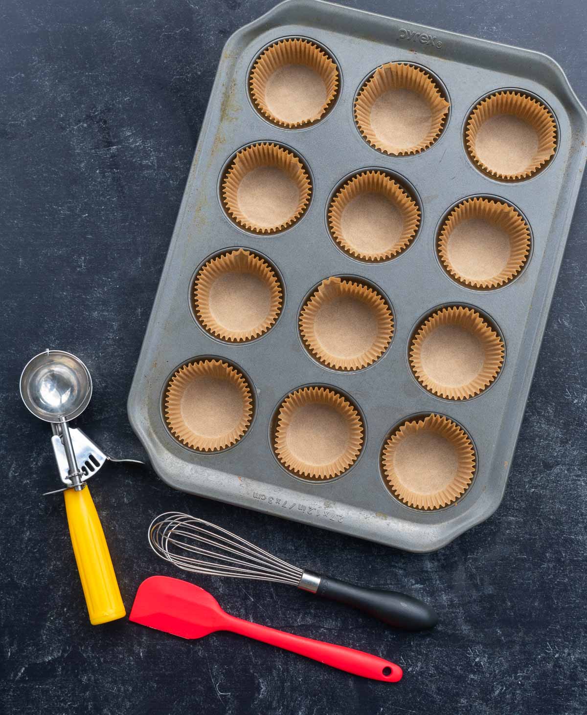 Tools needed for sourdough corn muffins, including a muffin pan lined with muffin liners, a whisk, spatula, and an optional cookie scoop.