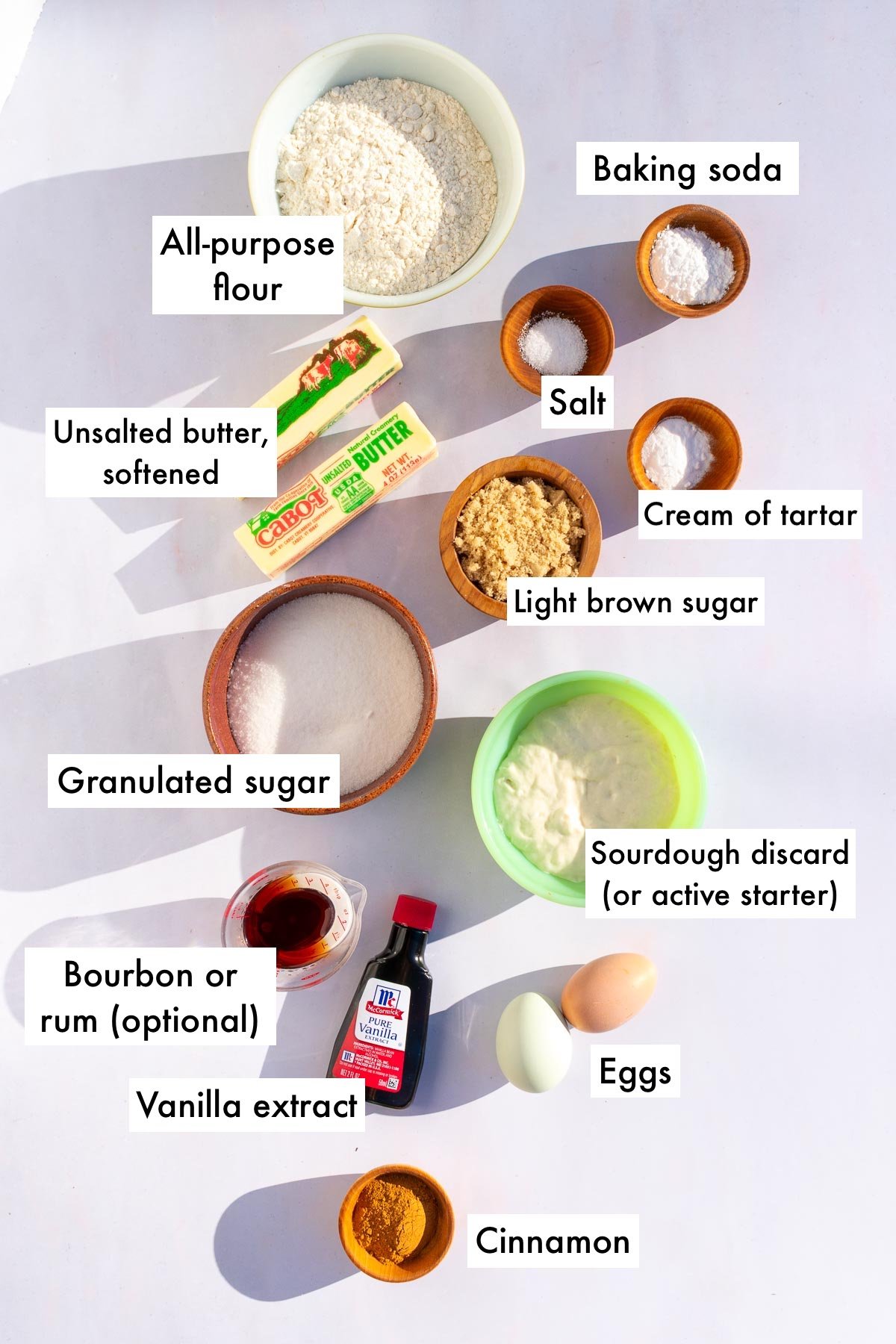 Labeled bowls of ingredients needed for sourdough snickerdoodle cookies including all-purpose flour, baking soda, unsalted butter, cream of tartar, salt, sourdough discard, vanilla extract, bourbon, cinnamon, light brown sugar, and granulated sugar.