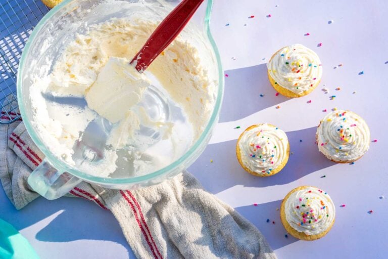 Sourdough frosting in a stand mixer bowl with a spatula and frosted cupcakes with rainbow sprinkles.