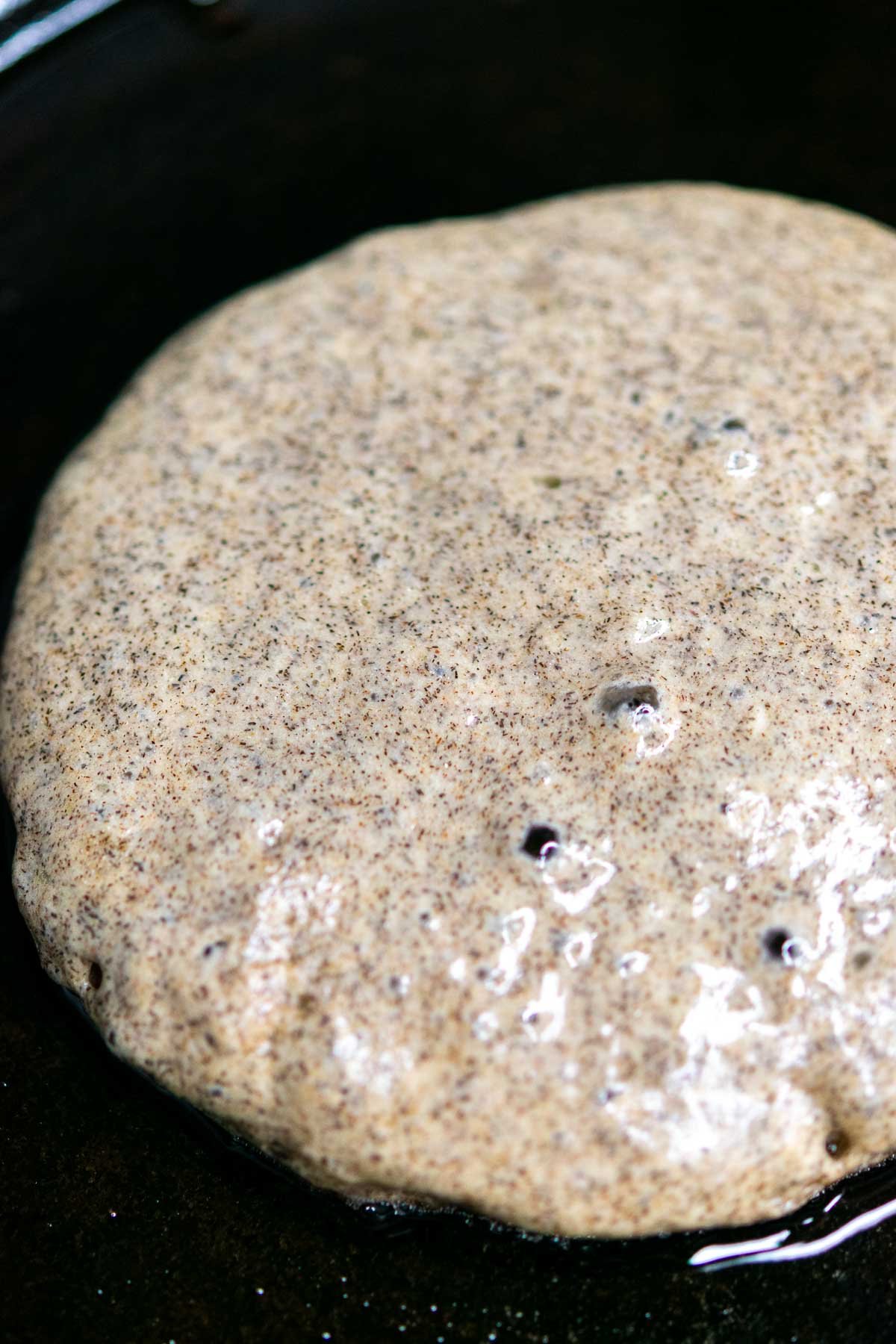A sourdough buckwheat pancake with bubbles in a cast iron skillet before flipping.