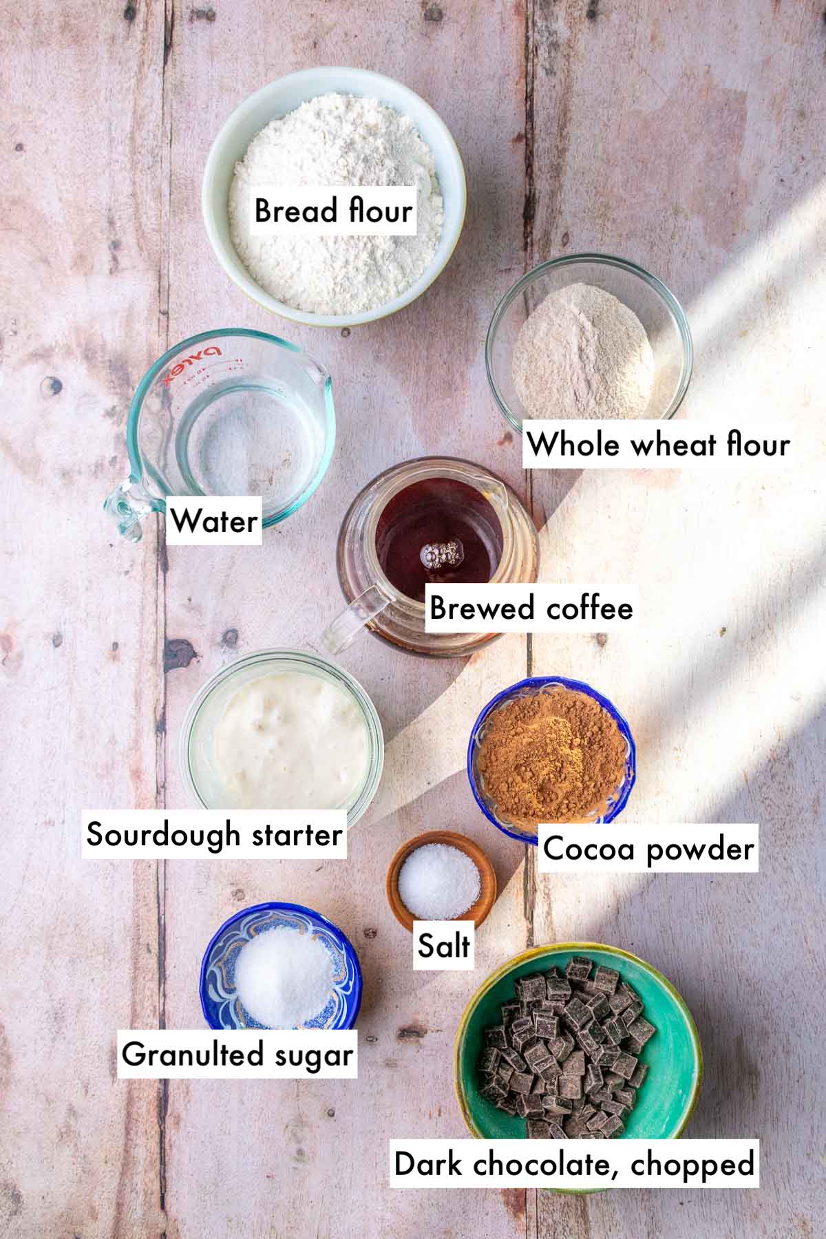 Bowls of labeled ingredients needed for chocolate coffee sourdough bread including bread flour, whole wheat flour, cocoa powder, chocolate, salt, water, sourdough starter, sugar, and brewed coffee.