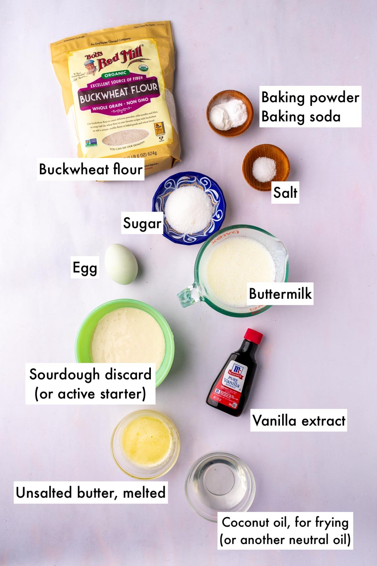 Labeled bowls of ingredients needed for sourdough buckwheat pancakes, including buckwheat flour, baking powder, baking soda, salt, sugar, buttermilk, sourdough discard, vanilla extract, melted butter, egg, and coconut oil.
