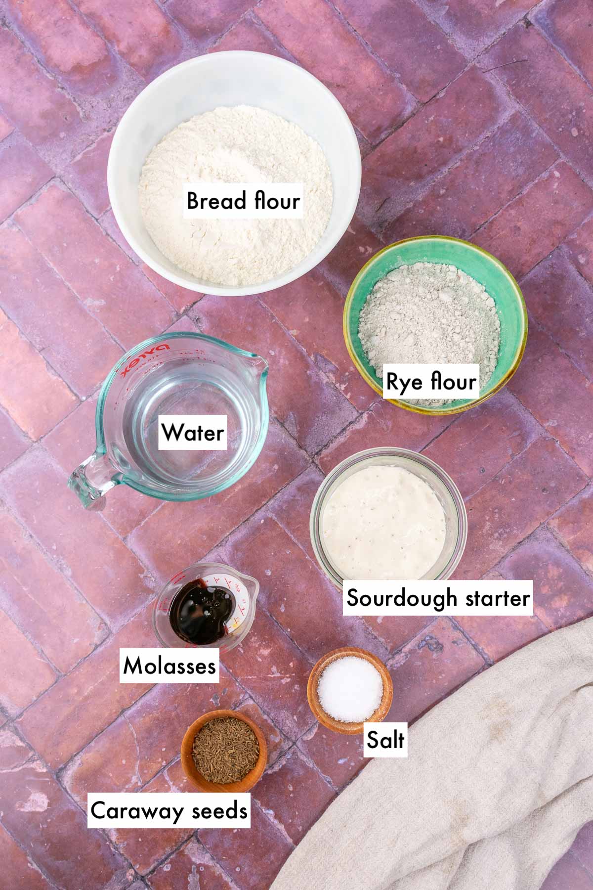 Labeled bowls of ingredients needed for sourdough rye sandwich bread, including bread flour, rye flour, water, molasses, salt, sourdough starter, and caraway seeds.