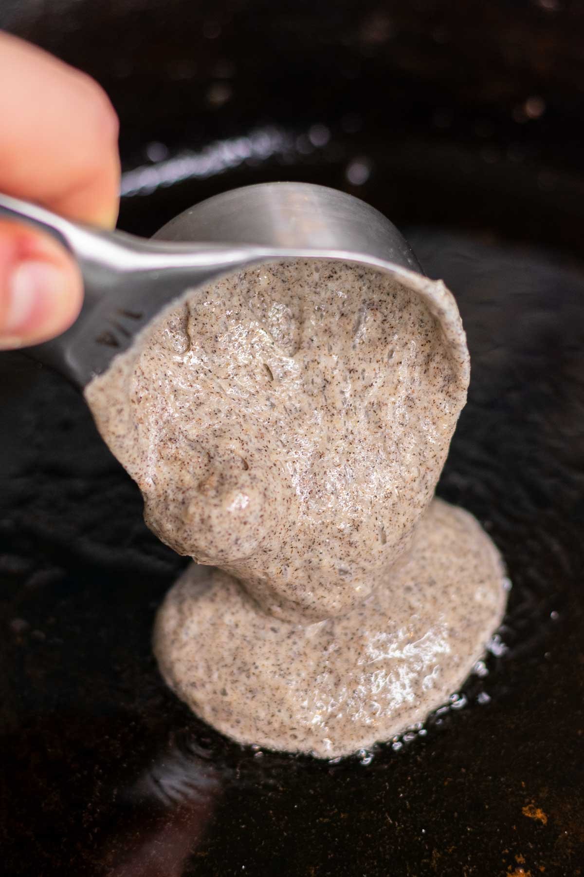 Scooping ¼ cup scoop of sourdough buckwheat pancake batter into a cast iron skillet with oil.