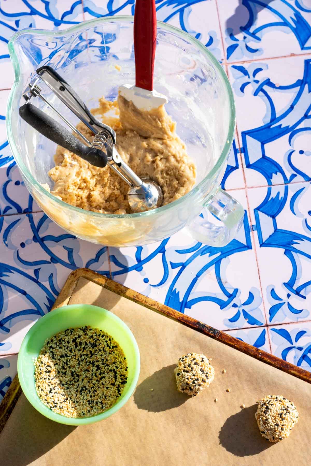 A mixing bowl with sourdough tahini honey cookies batter with a cookie scoop and spatula and a baking sheet with sesame seeds and portioned cookies.