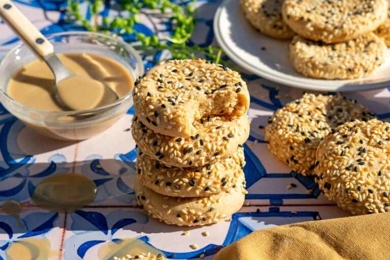 Stack of sourdough tahini honey cookies with a bite taken out of the top one and coated in white and black sesame seeds with a bowl of tahini in the background.