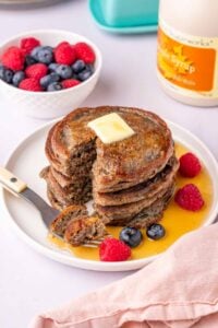 Stack of sourdough buckwheat pancakes with a fork taking a bite out and maple syrup, fruit, and butter.