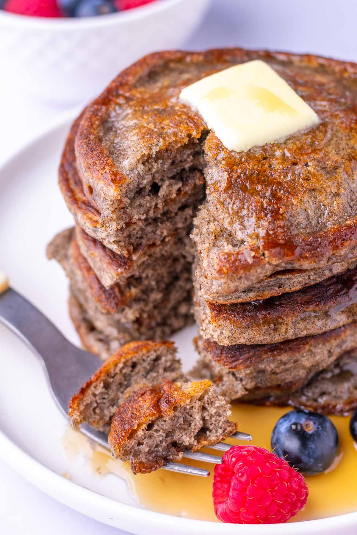 Stack of sourdough buckwheat pancakes with a fork bite taken out of them with syrup, fruit, and butter.