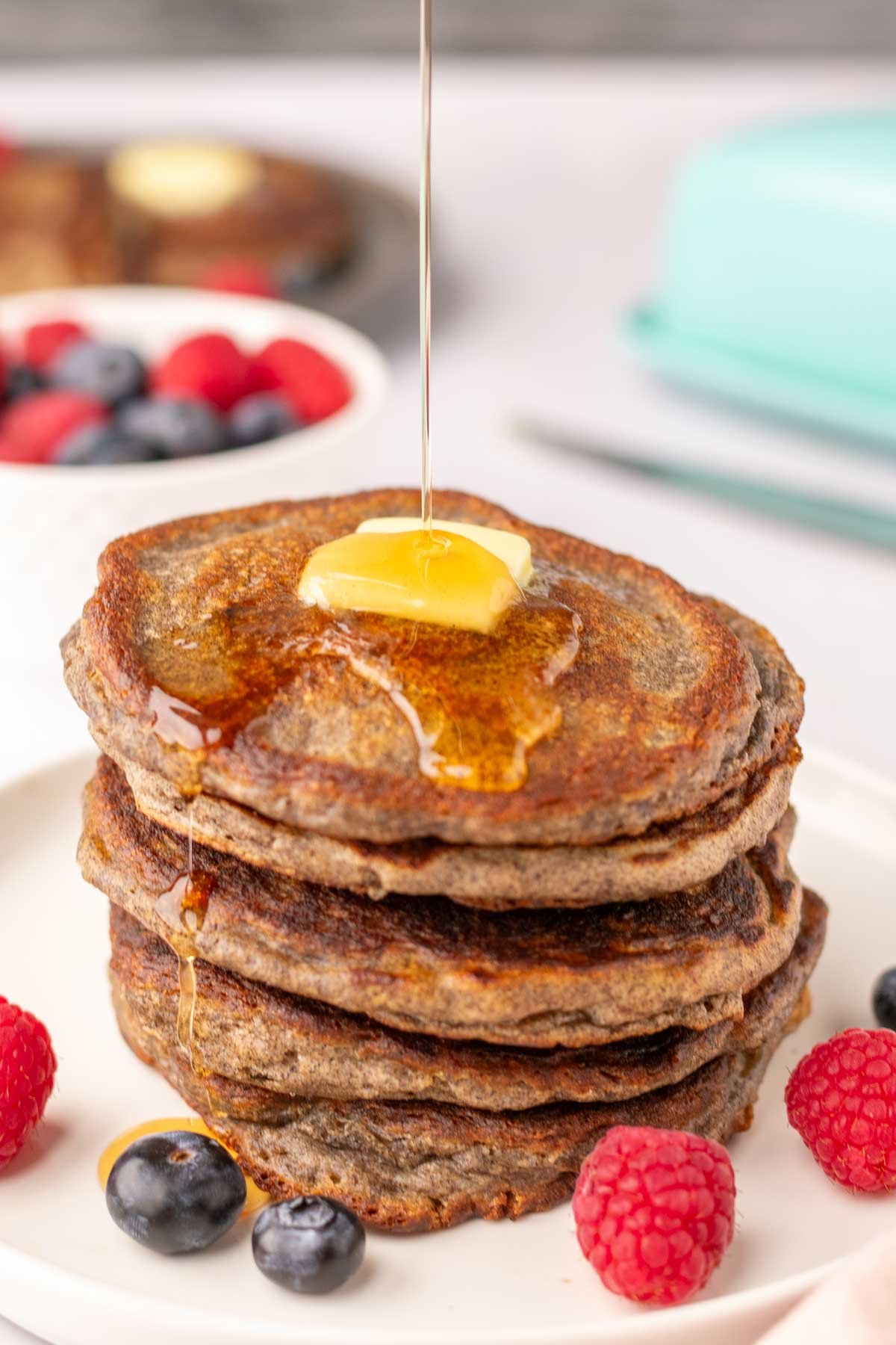Drizzling maple syrup on top of a stack of sourdough buckwheat pancakes with fruit and butter.