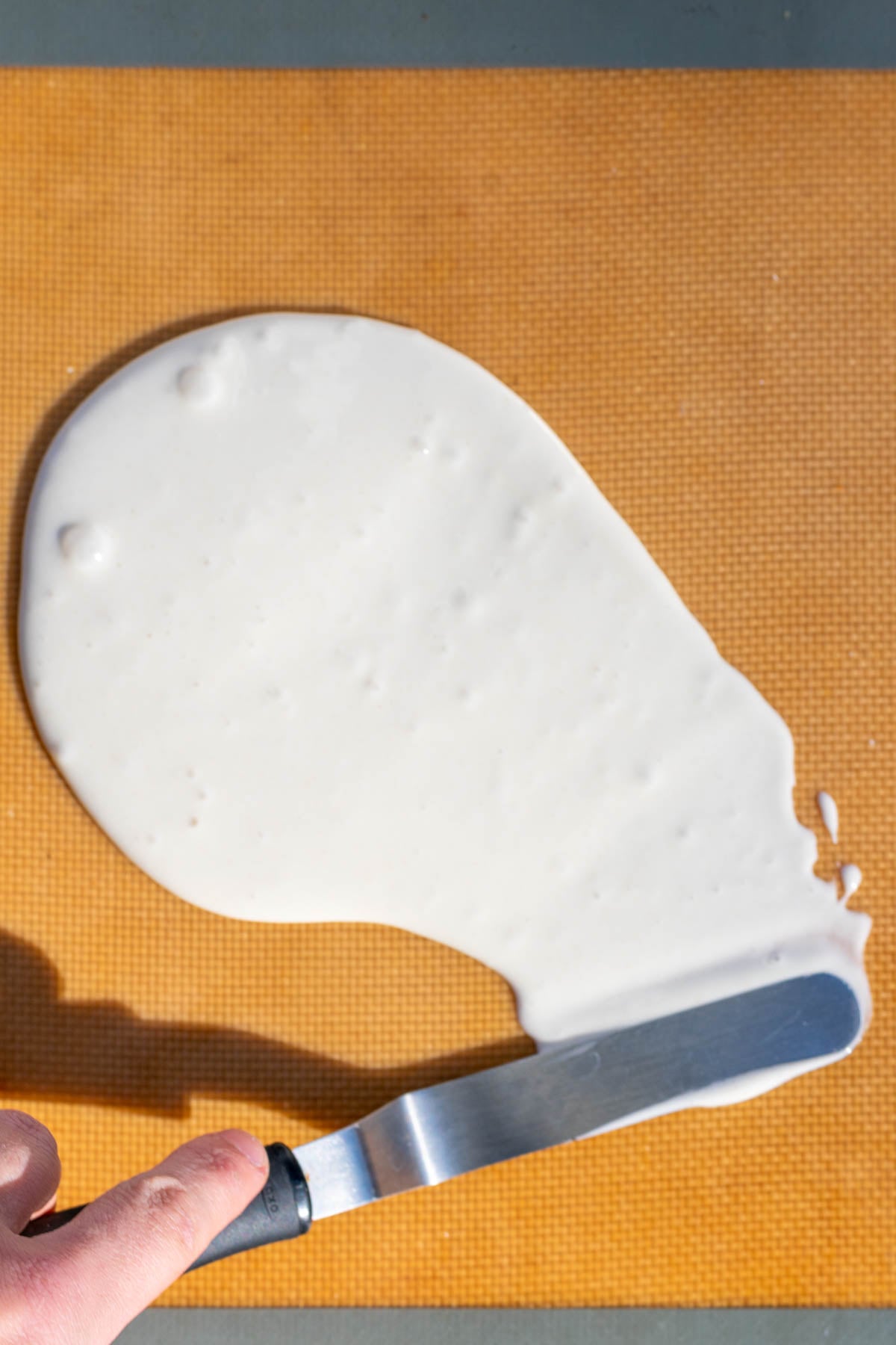 Hand spreading sourdough starter thinly on a silicone baking mat with an offset spatula.