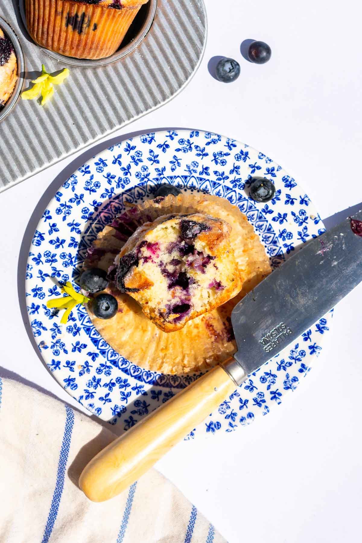 A bite taken out of a sourdough blueberry muffin on a plate with a knife and fresh blueberries.
