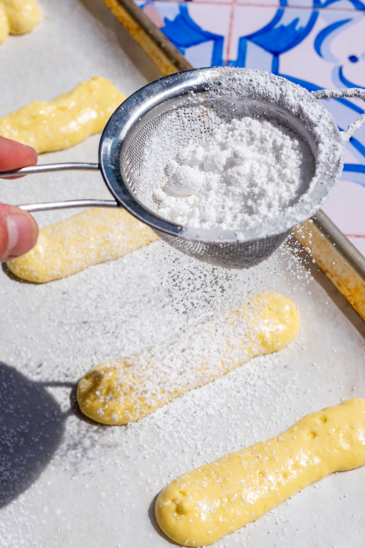 Dusting piped sourdough ladyfingers on a baking sheet with powdered sugar.