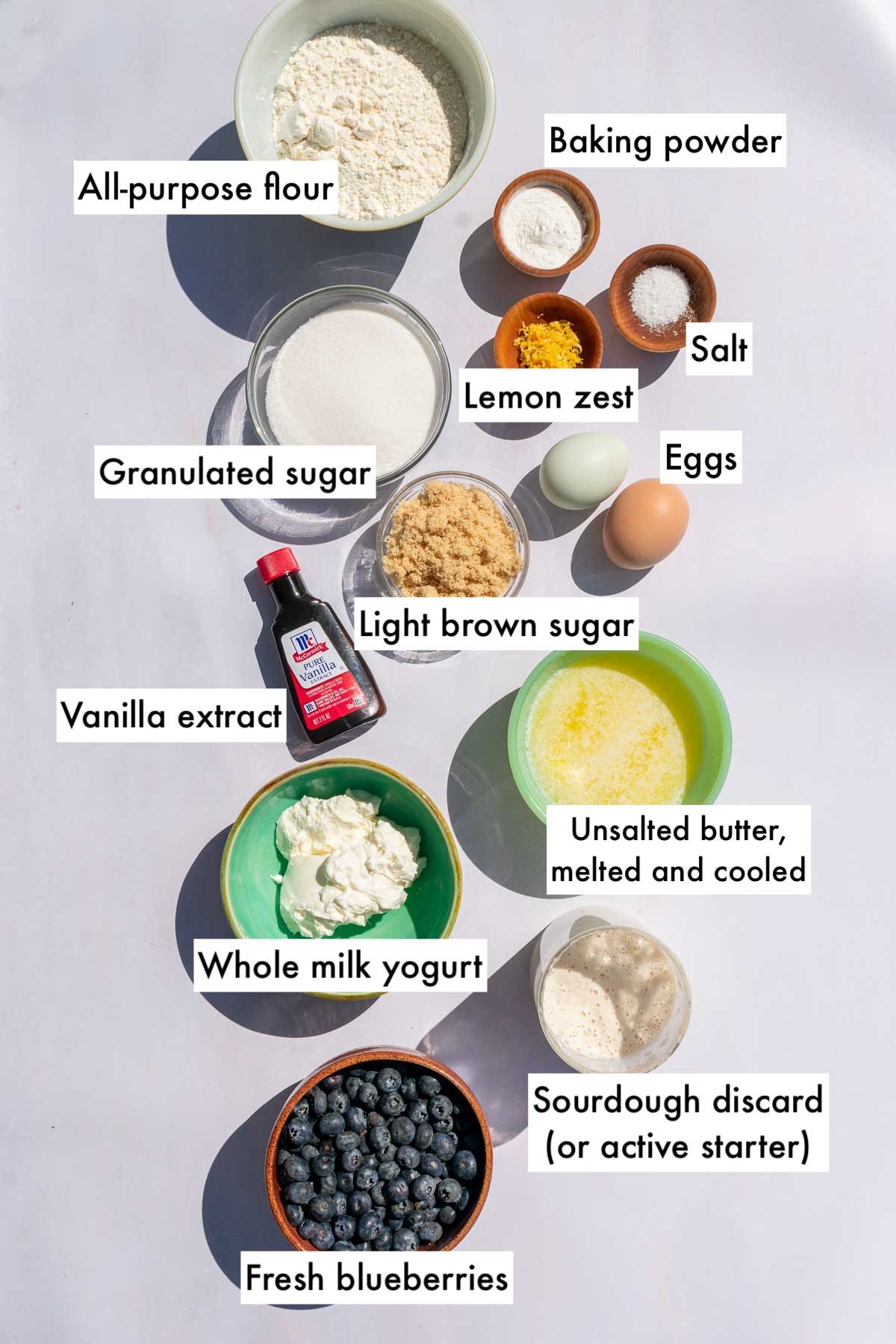Labeled bowls of ingredients needed for sourdough blueberry muffins including all-purpose flour, baking powder, salt, granulatd sugar, light brown sugar, eggs, vanilla extract, Greek yogurt, melted butter, fresh blueberries, and sourdough discard.