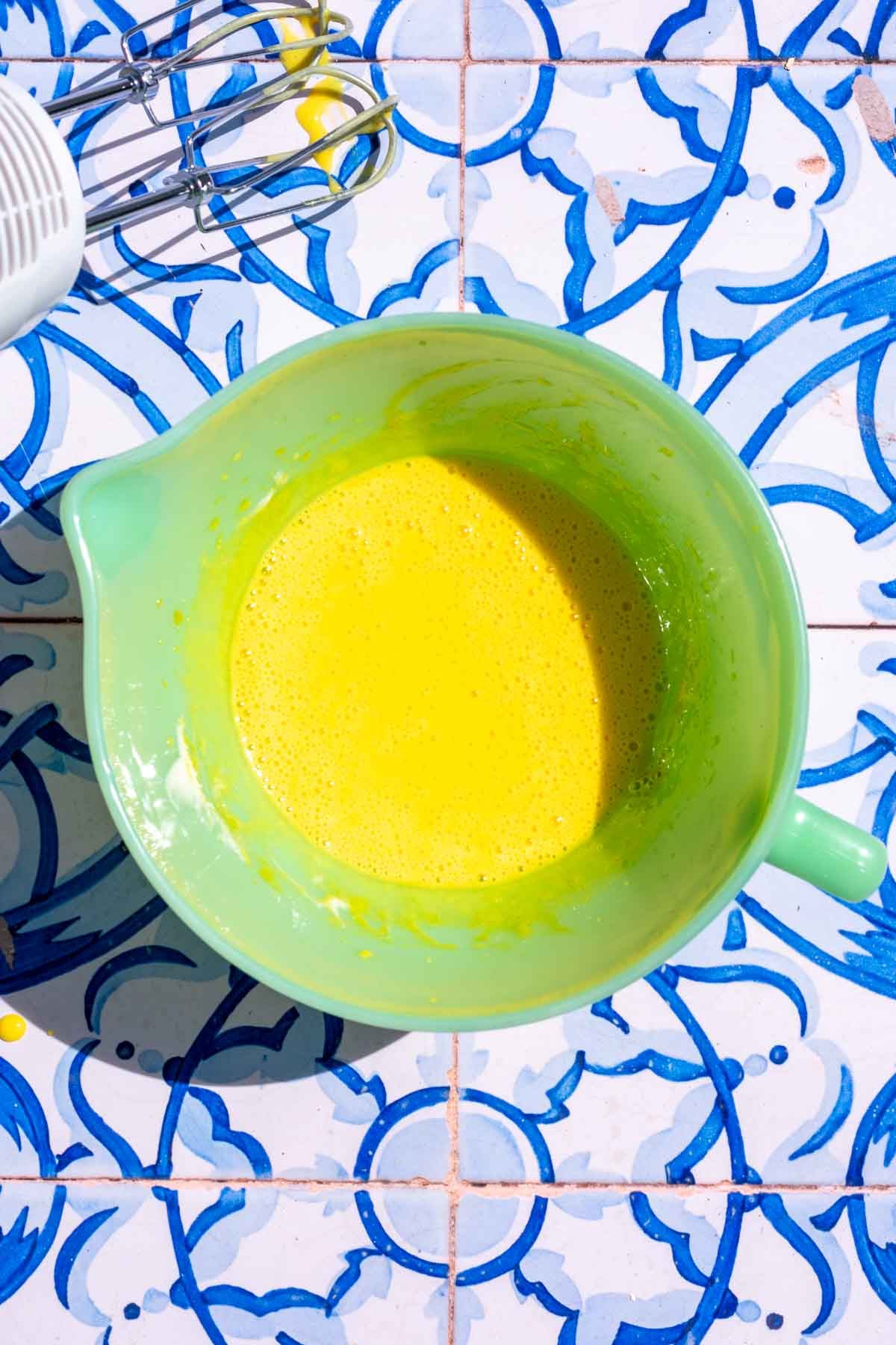 Mixed egg yolk mixture for ladyfingers in a green liquid bowl with an electric hand mixer to the side.