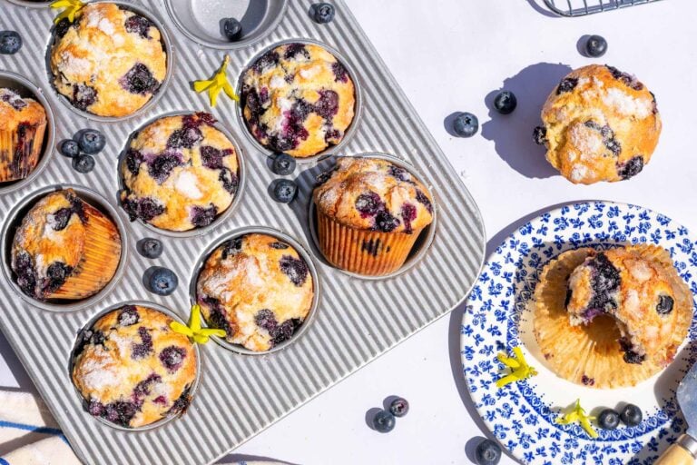 Sourdough blueberry muffins in a muffin pan and one with a bite taken out of it on a plate with fresh blueberries scattered around.