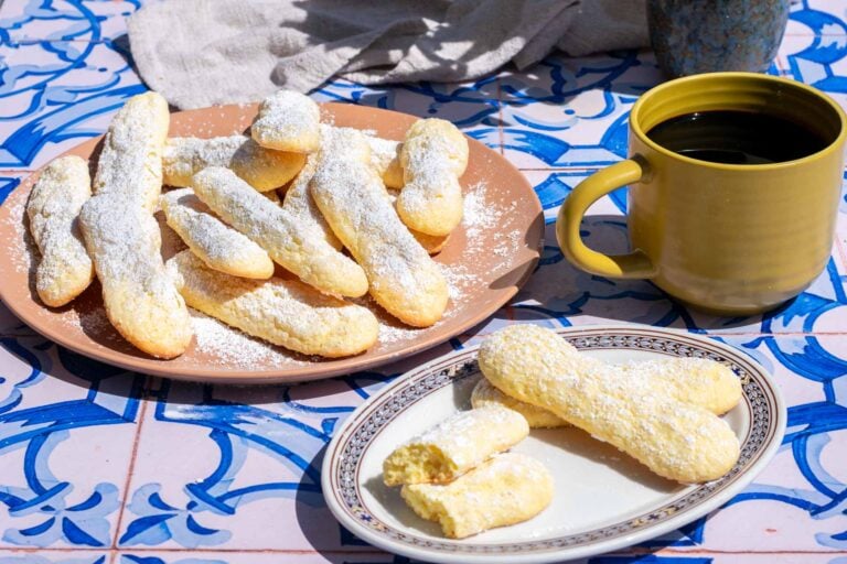 Sourdough ladyfingers dusted with powdered sugar on plates with a mug of coffee.